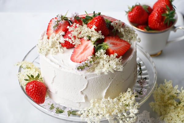 Pale pink layer cake topped with fresh strawberries and elderflower blossom